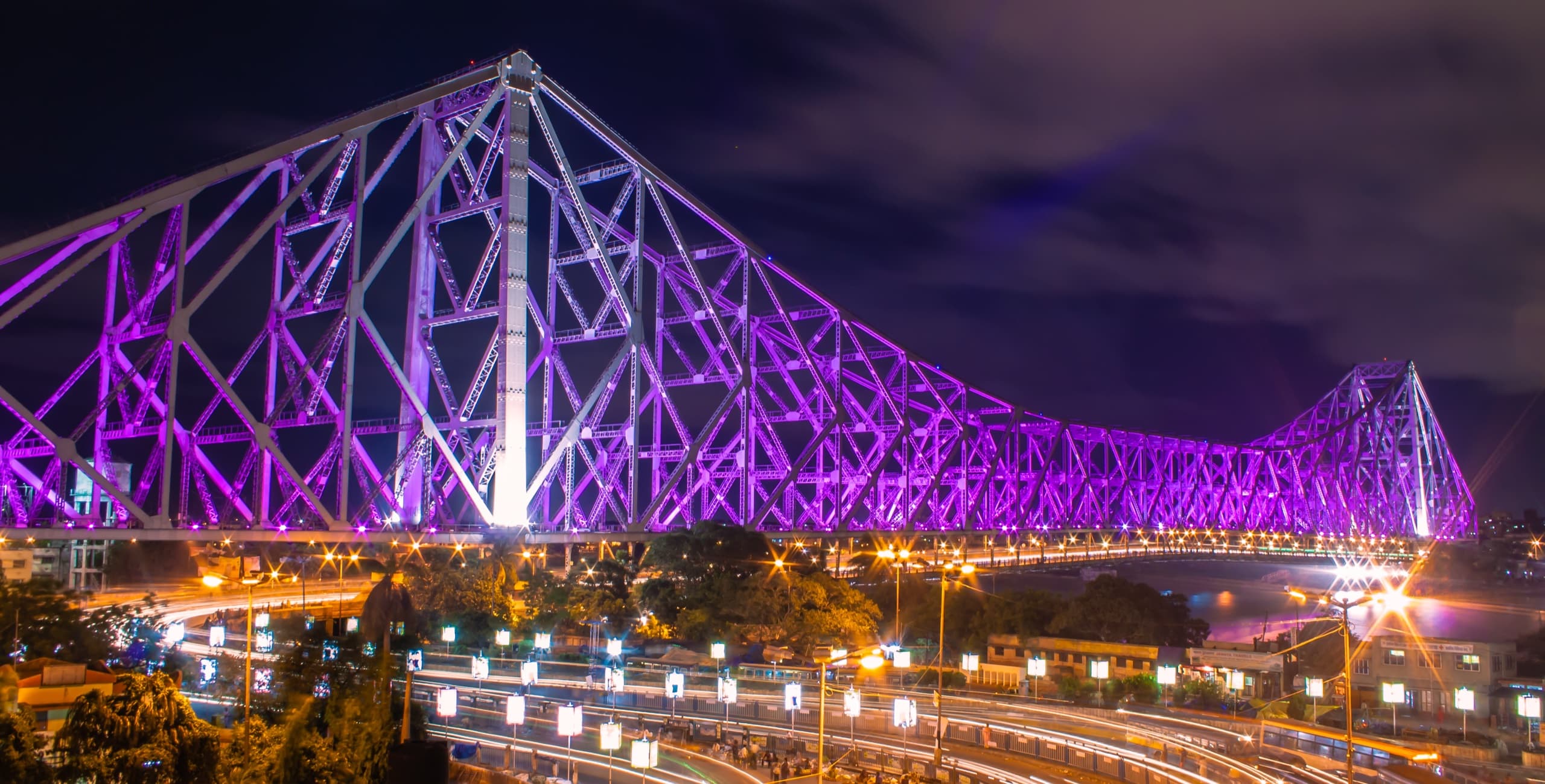 Howrah Bridge lit purple at night, above the light trails and street lamps of Bankim Setu